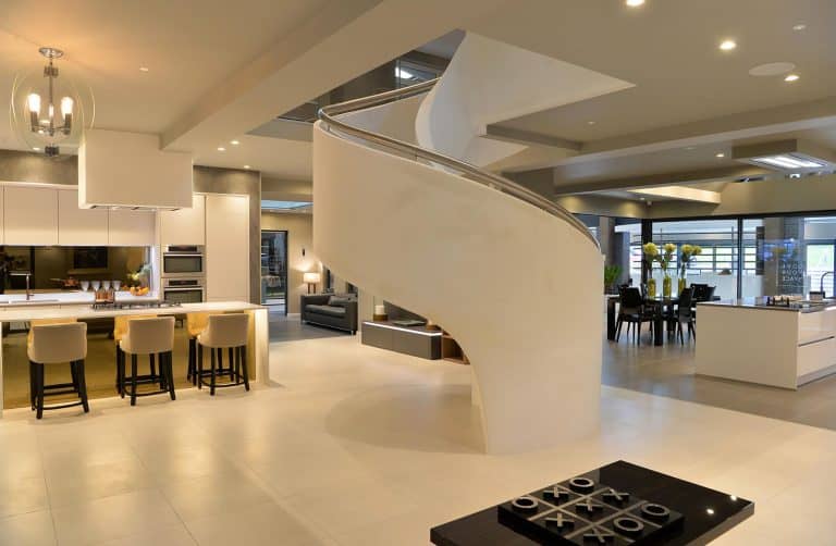 Modern, spacious open-plan living area with a central white spiral staircase, large kitchen island with bar stools, dining table, and bright lighting. Neutral tones and sleek, contemporary furniture throughout.