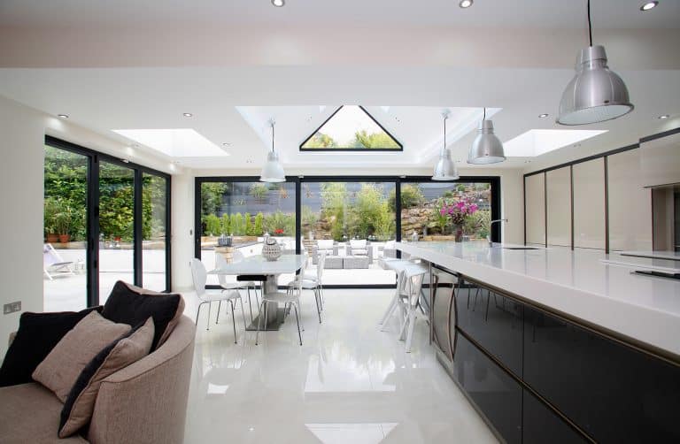 Modern, spacious kitchen and dining area with white floors, sleek countertops, pendant lights, glass doors, and large windows overlooking a bright patio and lush garden. A beige sofa sits in the foreground.