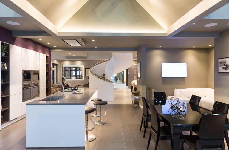 Modern open-concept kitchen and living area with a large island, black dining table, white sofa, wall-mounted TV, and a spiral staircase leading to an upper floor under vaulted ceilings with recessed lighting.