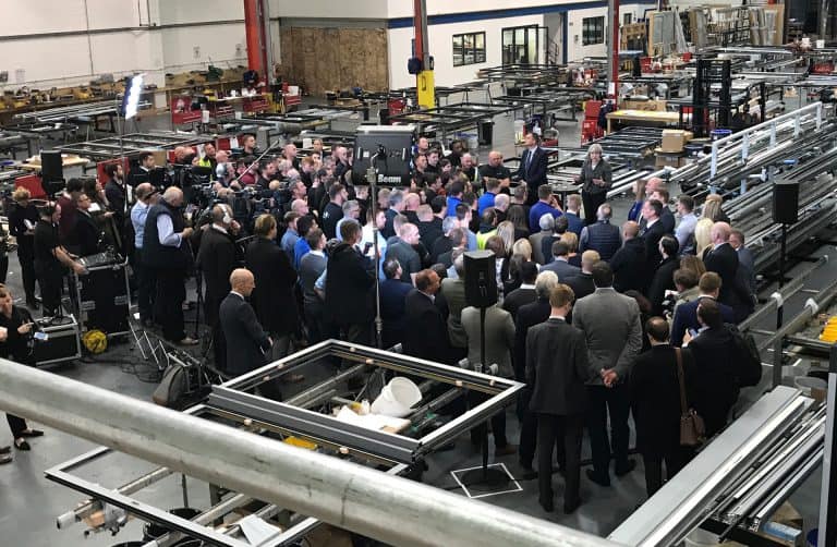 A large group of people, including media and camera crews, gather around speakers in a busy industrial factory with machinery, metal frames, and workstations visible in the background.