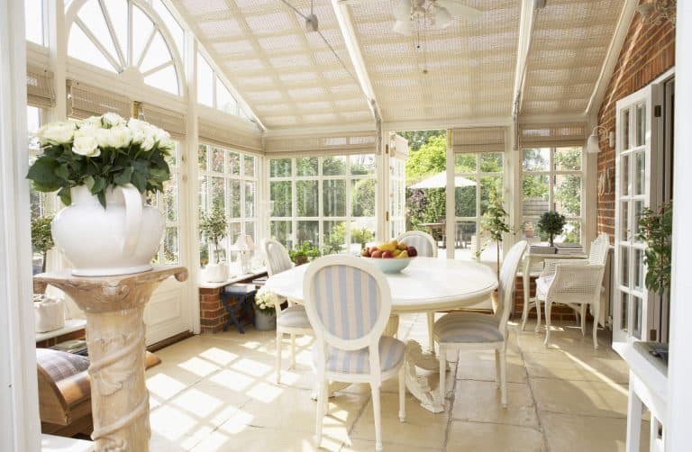 Bright sunroom with large windows, white furniture, striped chairs, a round dining table with fruit bowl, potted plants, and a vase of white roses on a pedestal. Sunlight streams through the glass roof.