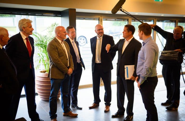 A group of six men in business attire stand in conversation indoors, while a sound crew records them using a boom microphone. One man gestures while holding a folder. Large windows and a potted plant are visible in the background.