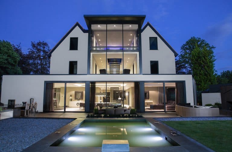 Modern three-story house with large glass windows and balconies, illuminated at dusk, featuring an open living area and a rectangular pool in the landscaped backyard.
