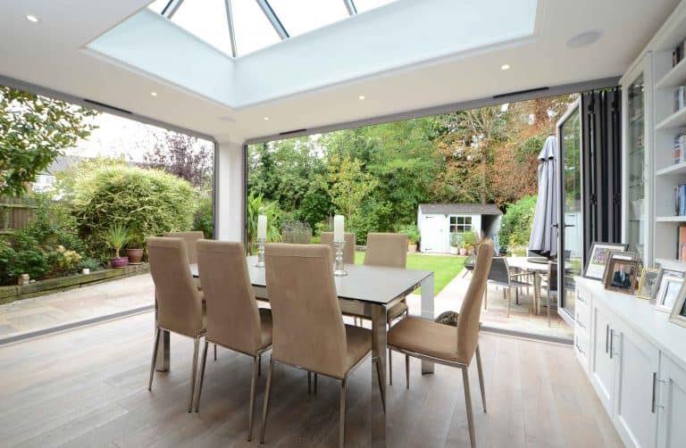 Modern dining room with beige chairs around a dining table, large skylight, and glass doors open to a lush green garden with a patio, tall plants, and a small white shed in the background.
