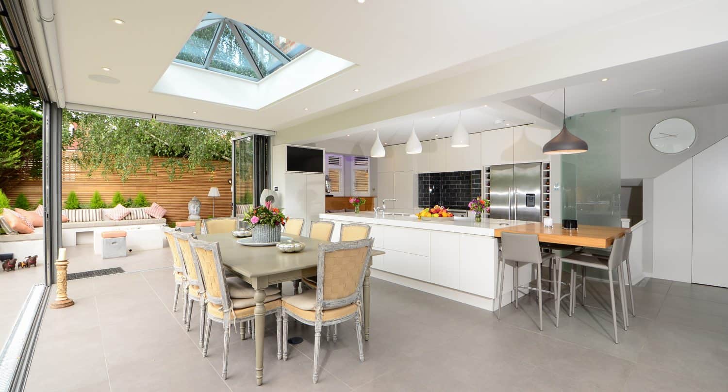 Modern open-plan kitchen and dining area with large skylight, sliding glass doors opening to a patio, sleek white cabinets, wooden accents, and neutral-toned furniture. Natural light fills the bright, spacious room.