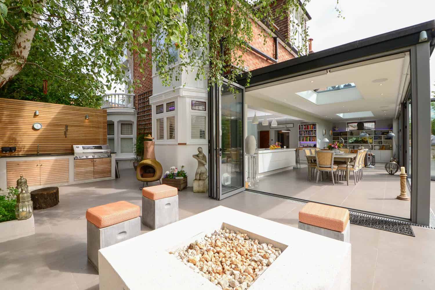 A modern outdoor patio with a fire pit, seating, a barbecue grill, and a wood wall. Sliding glass doors open to a bright kitchen and dining area, with large trees providing shade.