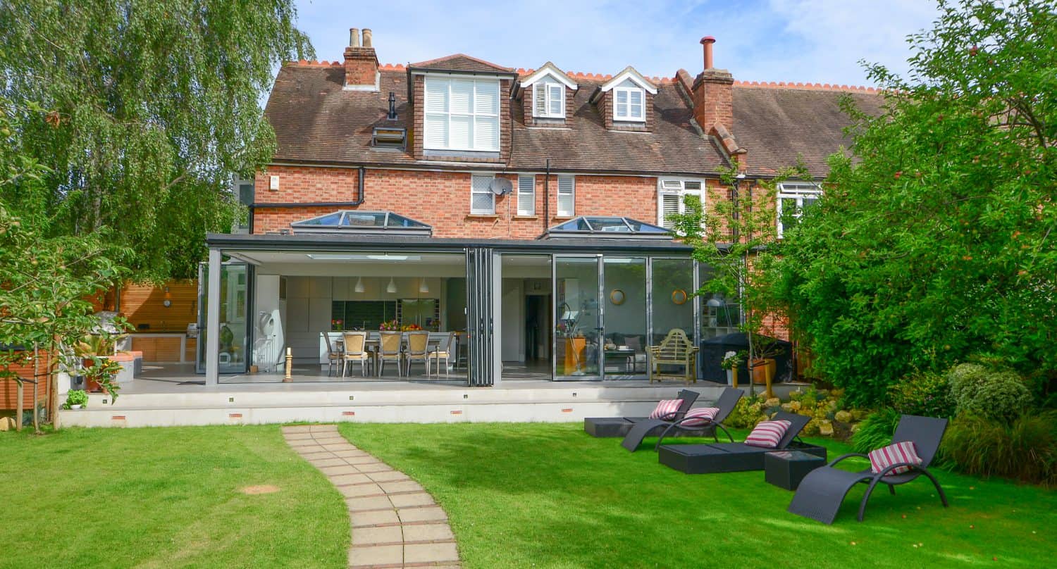 A modern brick house with glass roofing and a sleek glass extension opens onto a patio with outdoor dining. A path crosses a green lawn toward black lounge chairs with striped cushions, surrounded by trees and garden plants.