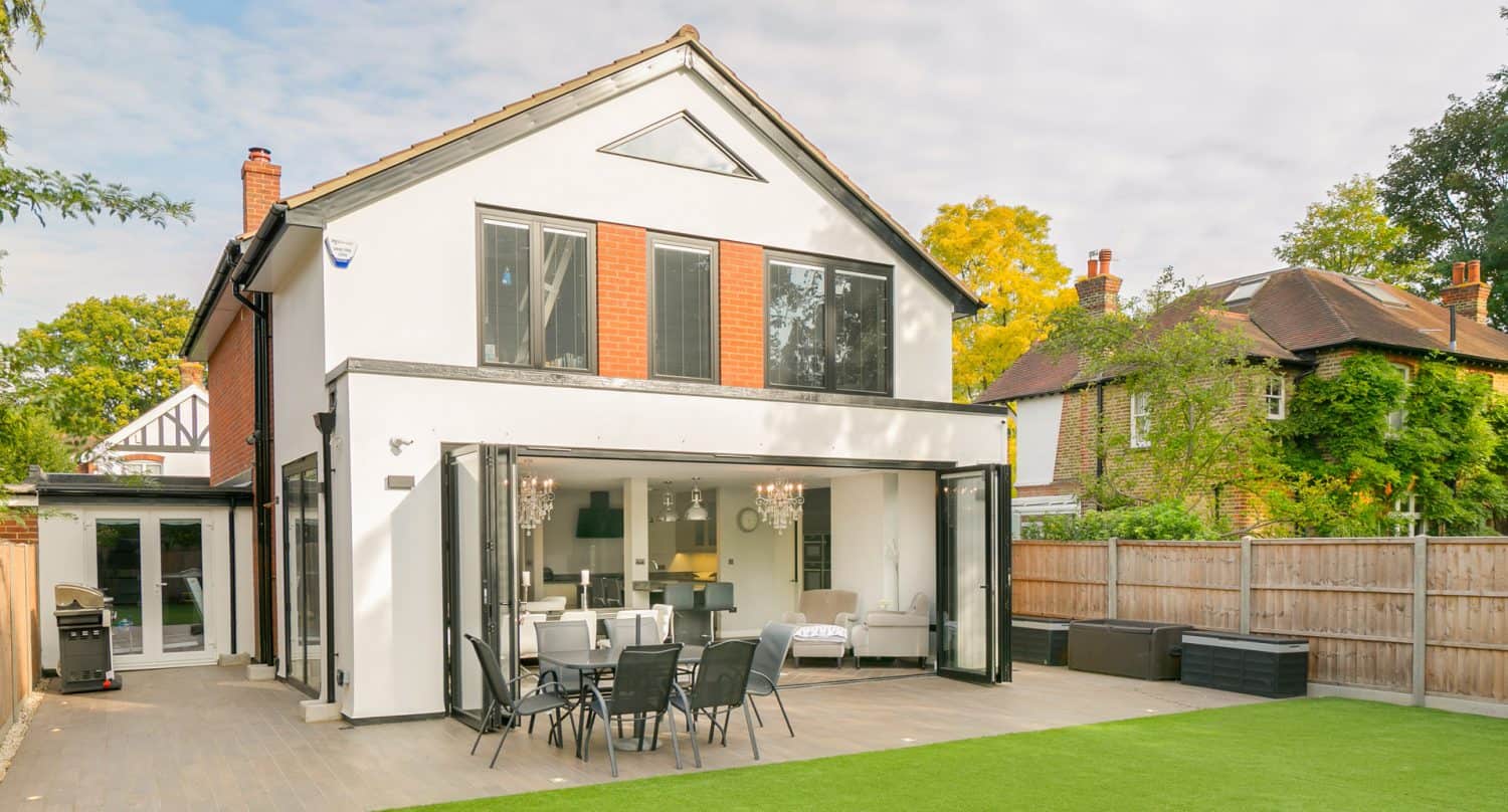 A modern two-story house with large glass doors open to a patio, showing an outdoor dining area and a spacious lawn. The house has red and white walls, and theres another house visible next door.