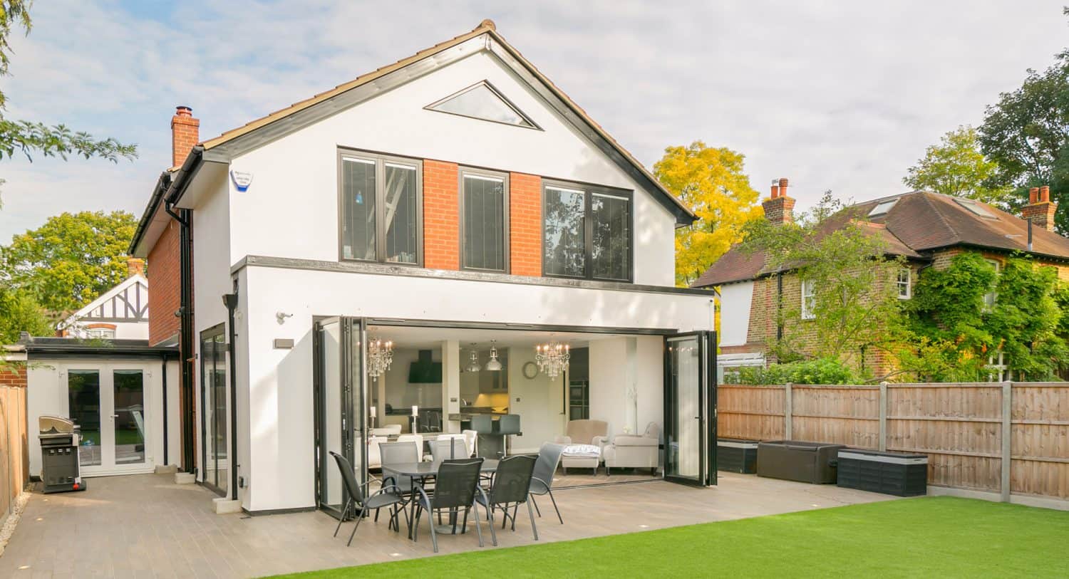 A modern two-story house with large glass doors open to a patio, showing an outdoor dining area and a spacious lawn. The house has red and white walls, and theres another house visible next door.