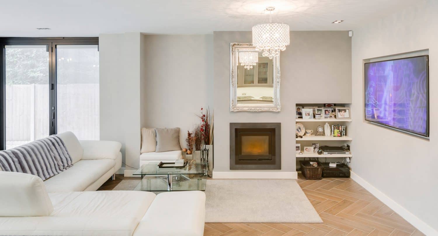 Modern living room with white sofas, a glass coffee table, a fireplace under a large ornate mirror, a wall-mounted TV, shelves with books and decor, wooden herringbone floor, and a chandelier.