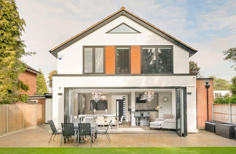 A modern two-story house with large windows and folding glass doors opening to a patio with outdoor dining furniture, surrounded by trees and a fenced yard.
