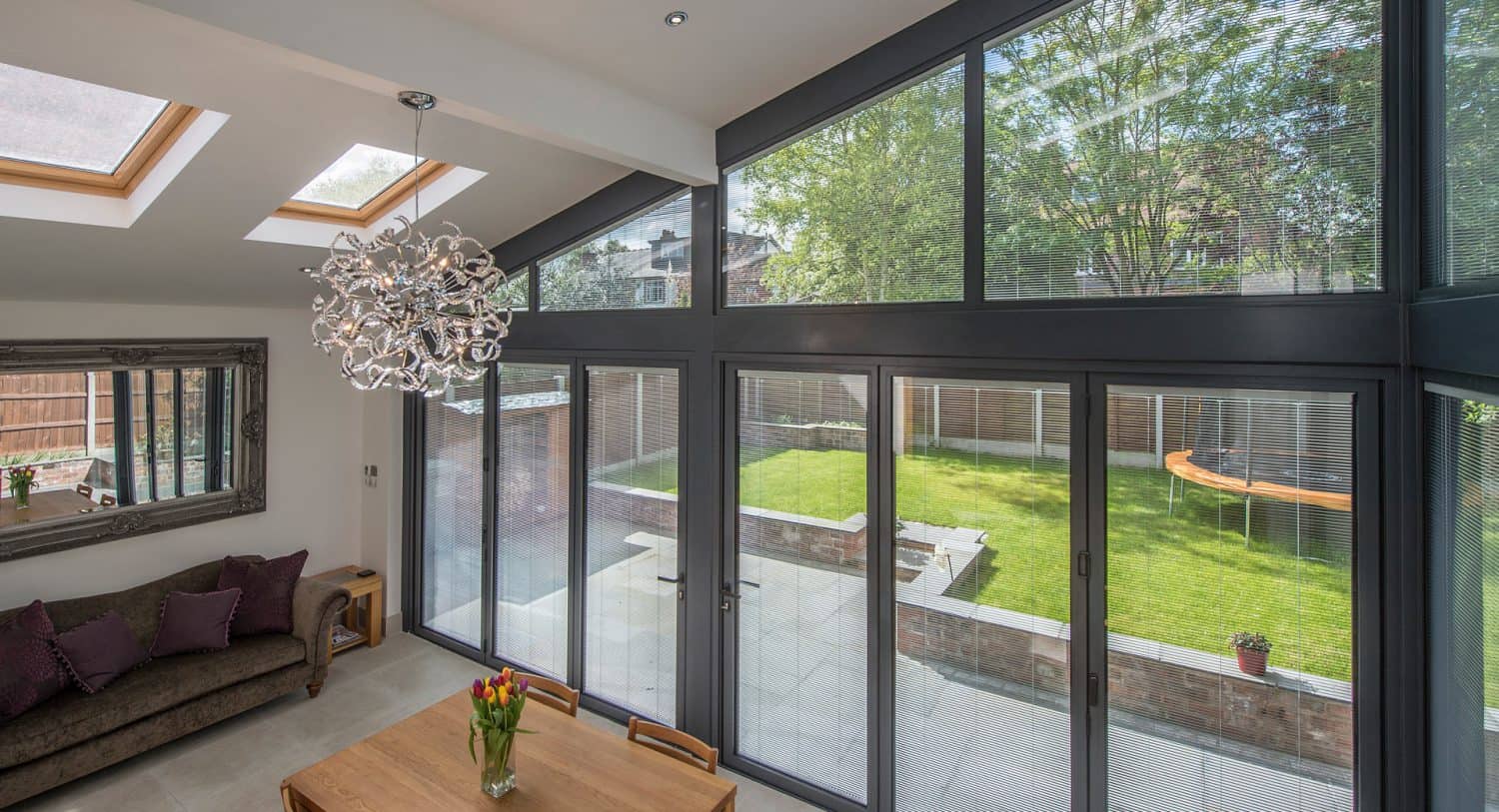 Modern sunroom with large glass doors and windows, a chandelier, a sofa with cushions, a wooden dining table with flowers, and views of a bright green backyard with trees and a trampoline.