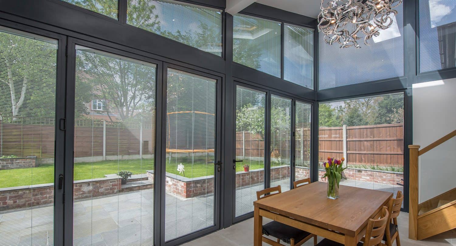 A modern dining area with large glass walls and doors, a wooden table with chairs, a chandelier, and a view of a green garden and patio outside. Natural light fills the bright, spacious room.