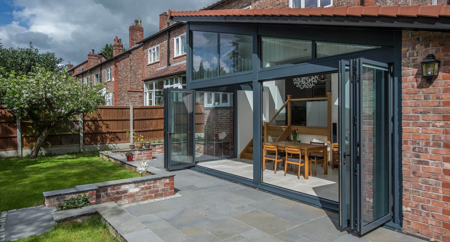 Modern brick house with large folding glass doors open to a patio and garden. The interior dining area with wooden table and chairs is visible, and there’s a green lawn with a tree and wooden fence outside.
