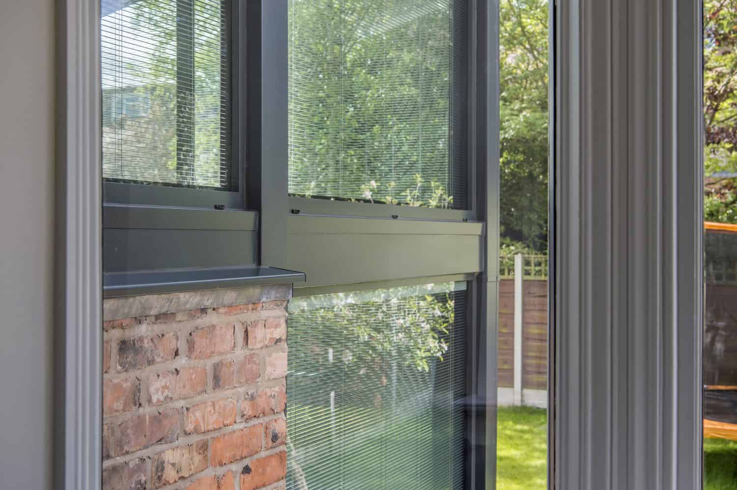 View from inside a building showing a large window with green metal frames, a section of exposed brick wall, and bifold doors opening to a garden with trees and grass visible outside.