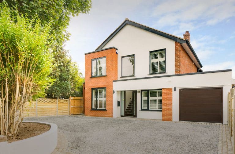 Modern two-story house with white and red brick exterior, large windows with shutters, a dark garage door, and a gravel driveway bordered by a wooden fence and landscaped greenery.