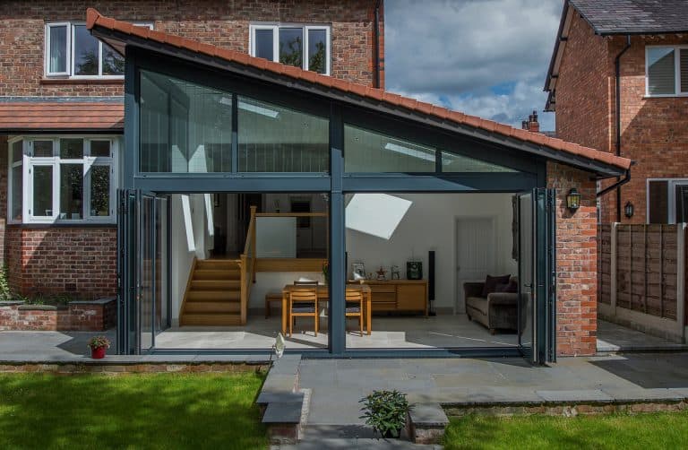 Modern glass extension on a brick house, featuring large folding doors, a slanted roof, visible wooden stairs, a small dining table, and a lounge area, opening onto a stone patio and green lawn.