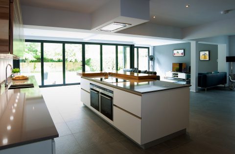 Modern open-plan kitchen with a large white island, built-in ovens, and sleek counters. Floor-to-ceiling glass doors open to a patio, allowing natural light to fill the spacious living and dining area.