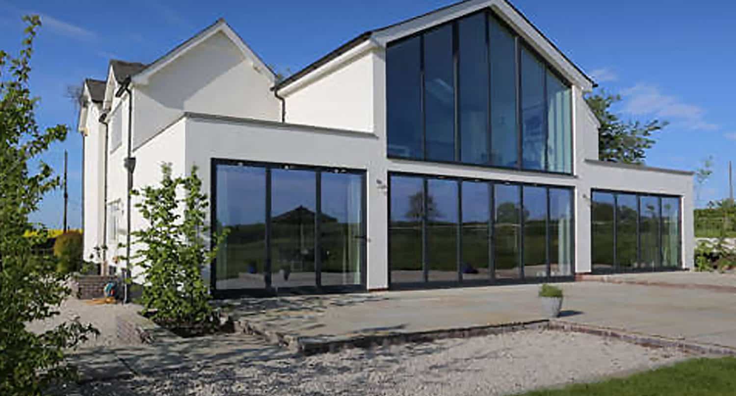 Modern white house with large floor-to-ceiling windows and a sloped roof, set against a clear blue sky. The house features an expansive patio area and is surrounded by greenery.