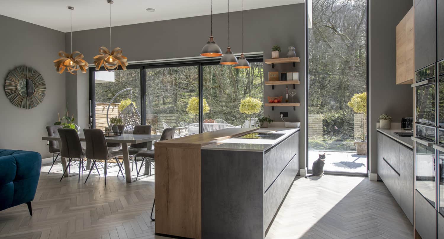 Modern kitchen and dining area with large bifold doors letting in sunlight. A black cat sits by the tall window. Neutral tones, wood, and dark accents decorate the space, with a view of a green garden outside.