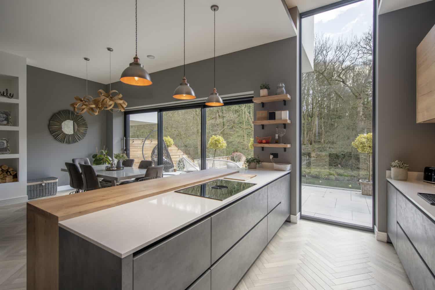 Modern kitchen with gray cabinets, a wooden countertop island, pendant lights, and large floor-to-ceiling bifold doors overlooking the patio and trees. Dining area with a round table is visible in the background.