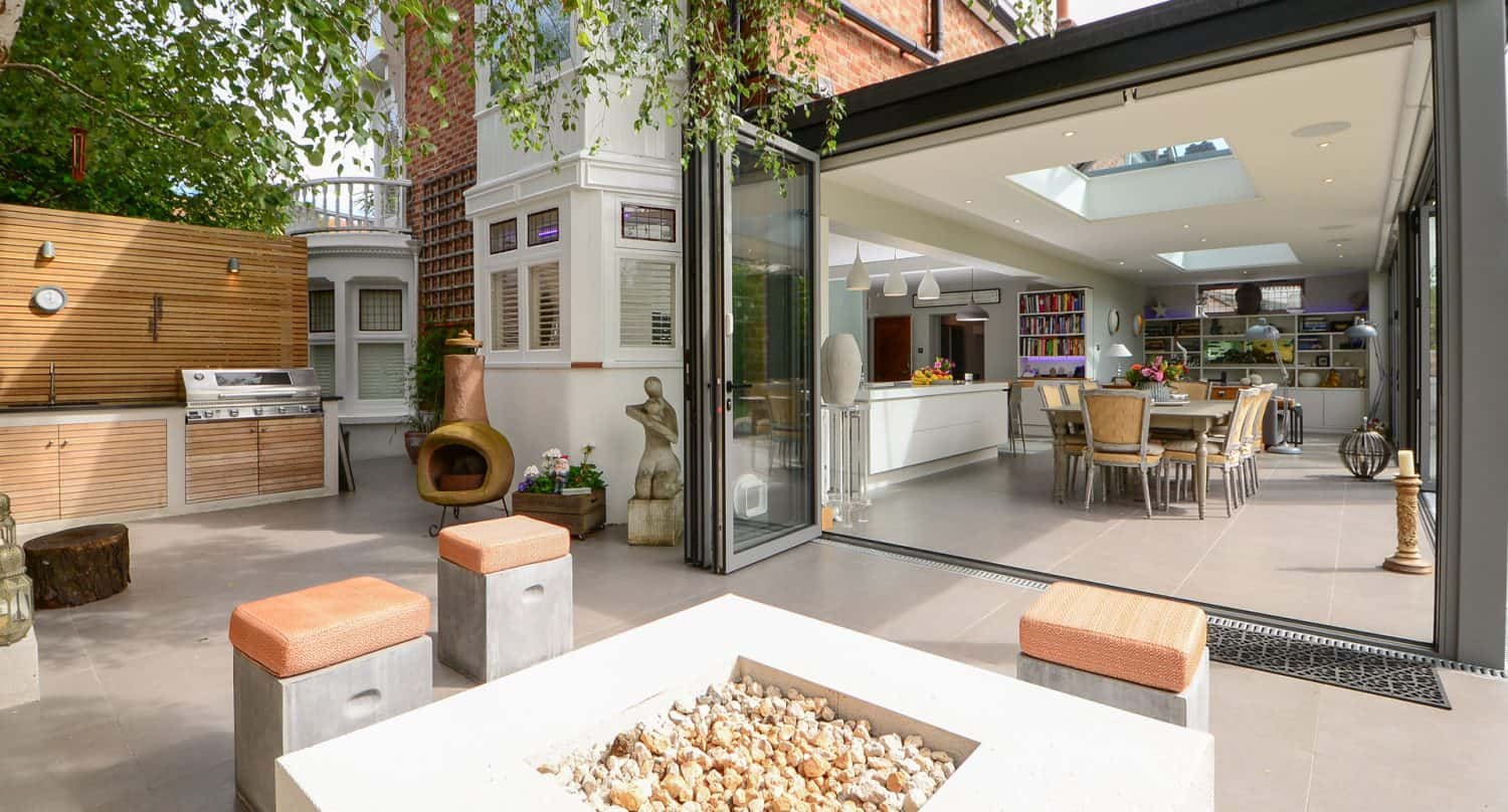 Modern open-plan kitchen and dining area with large glass doors opening to a stylish patio featuring a fire pit, outdoor seating, built-in grill, and greenery for a seamless indoor-outdoor living space.