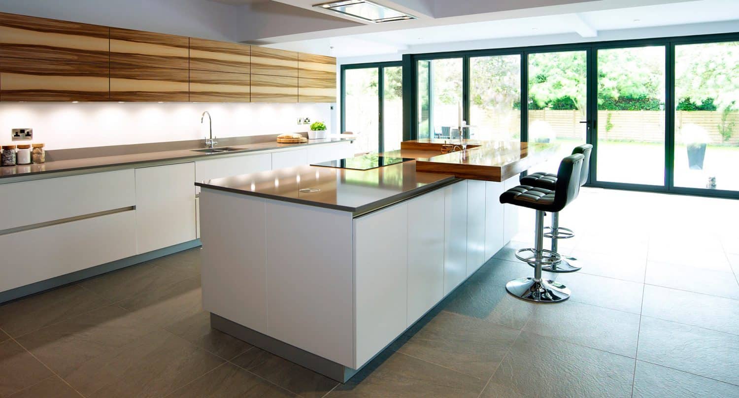 Modern kitchen with large windows, sleek white cabinets, wooden accents, a central island with two bar stools, and ample natural light coming from a garden view outside.