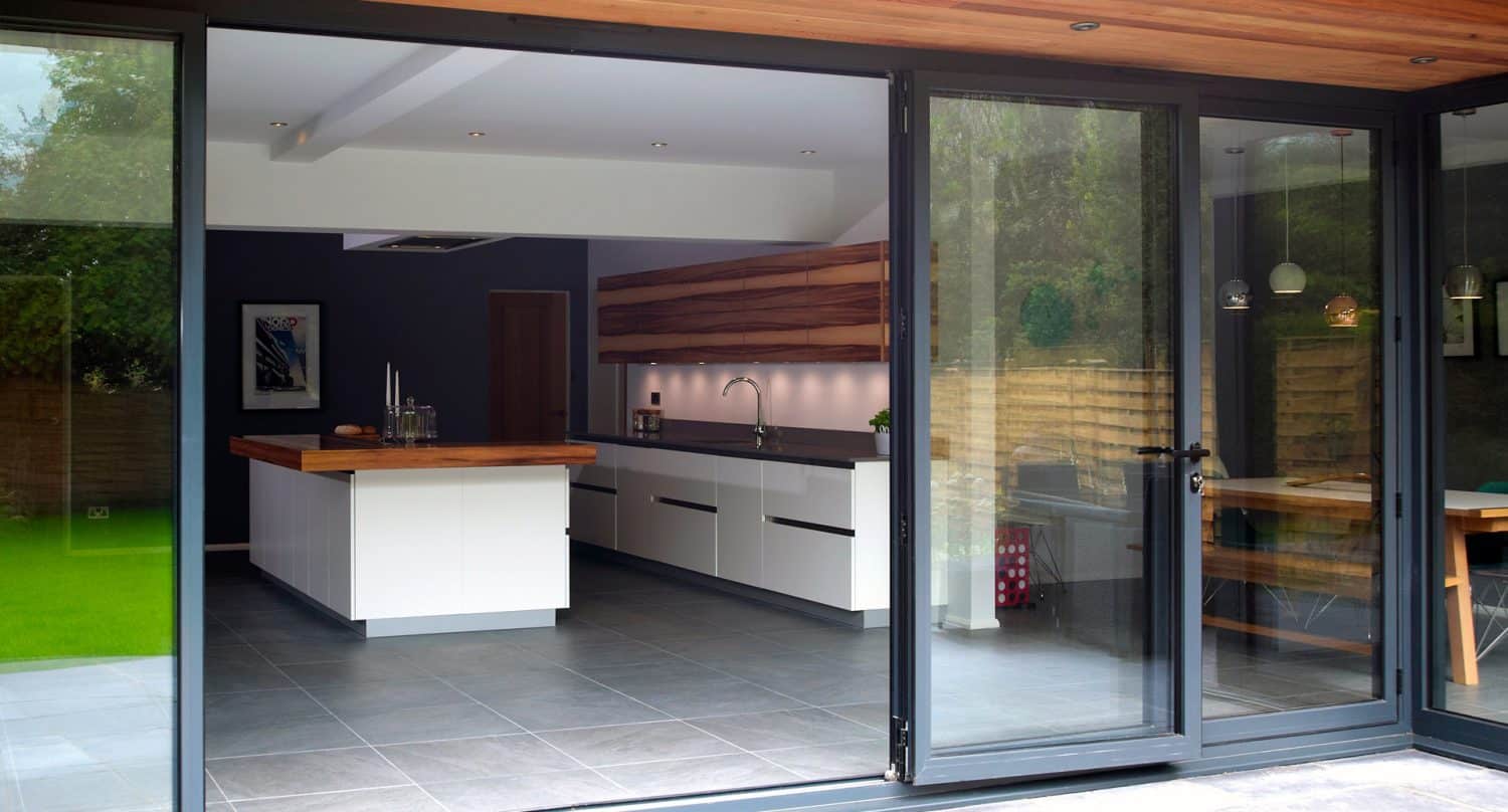 Modern kitchen with large glass sliding doors opening to a patio and garden, featuring white cabinets, a wooden island, grey tile floors, and minimalist decor. Natural light fills the space.