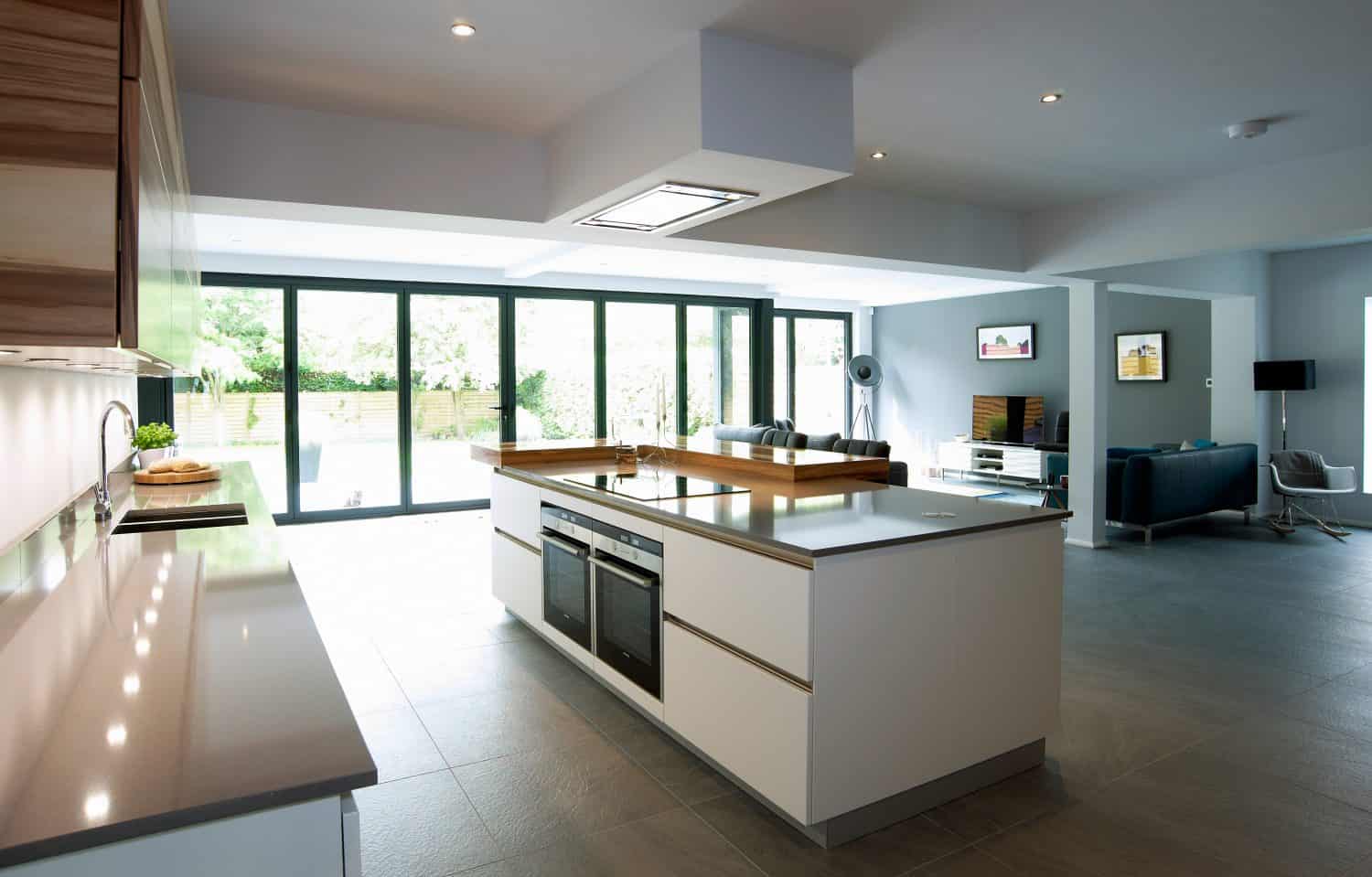 Modern, spacious kitchen with a large island, built-in oven, and minimalist white cabinetry. The open-plan room features bespoke glass solutions, including glass doors overlooking the garden, and connects to a bright living area with contemporary furniture.