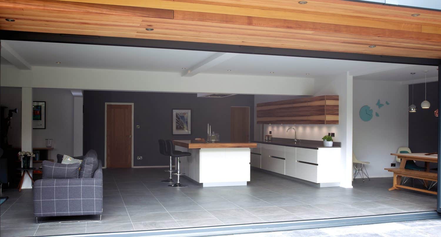 Modern open-plan kitchen and living area with large sliding doors and bespoke glass solutions, featuring a sofa, kitchen island with barstools, white cabinetry, wooden accents, a dining bench, and minimalist decor.