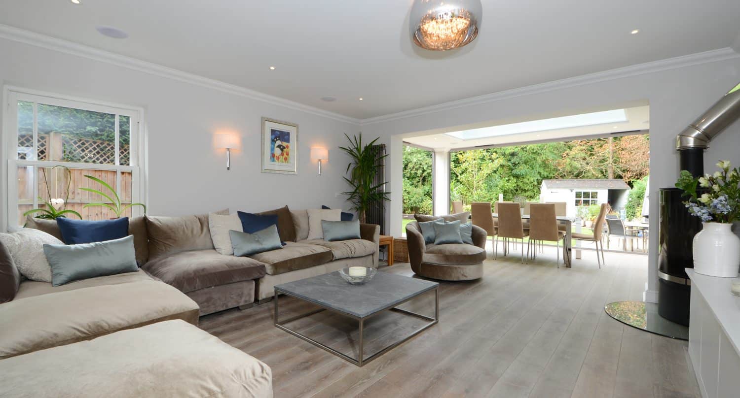 Modern living room with a large sectional sofa, coffee table, and decorative pillows; open dining area features bifold doors leading to a garden patio, with bright neutral tones and abundant natural light.