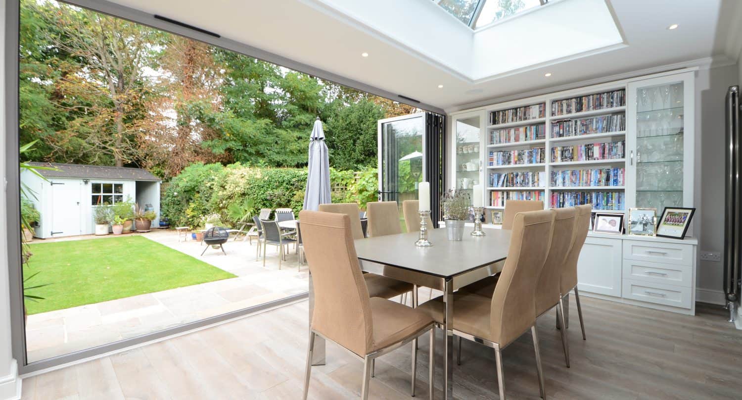 A modern dining room with a large table and beige chairs, featuring a glass ceiling and fully open bifold doors leading to a lush garden with a patio and small white garden shed. Shelves filled with books line one wall.