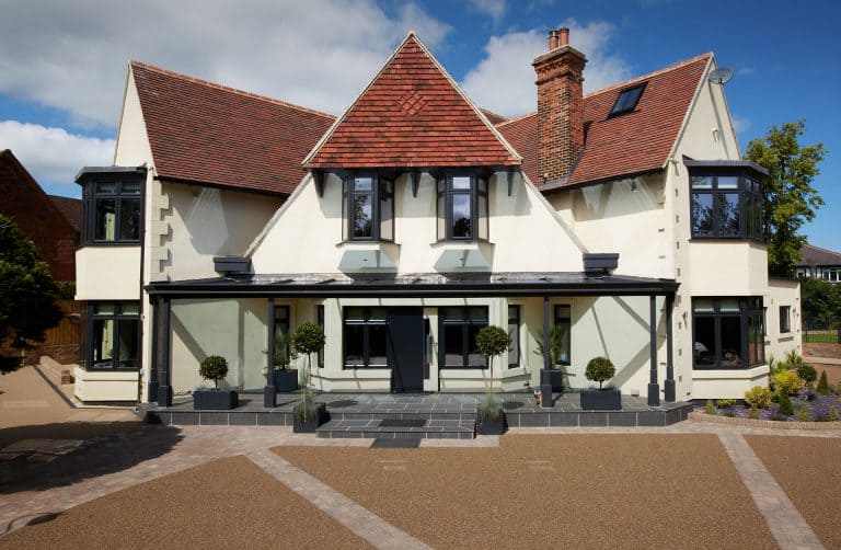 A large, modernized house with cream-colored walls, red-tiled roof, black window frames, and a covered front porch. The front yard features geometric landscaping and potted plants along the entrance.