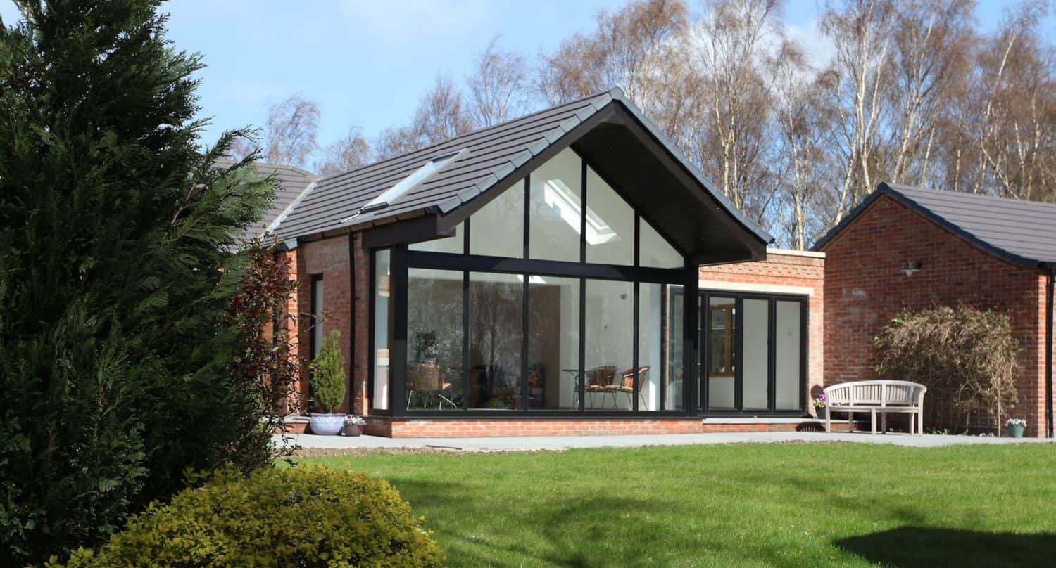 A modern brick house with a large glass extension featuring floor-to-ceiling windows, a sloped gray roof, and a patio with outdoor seating, surrounded by a green lawn and trees in the background.