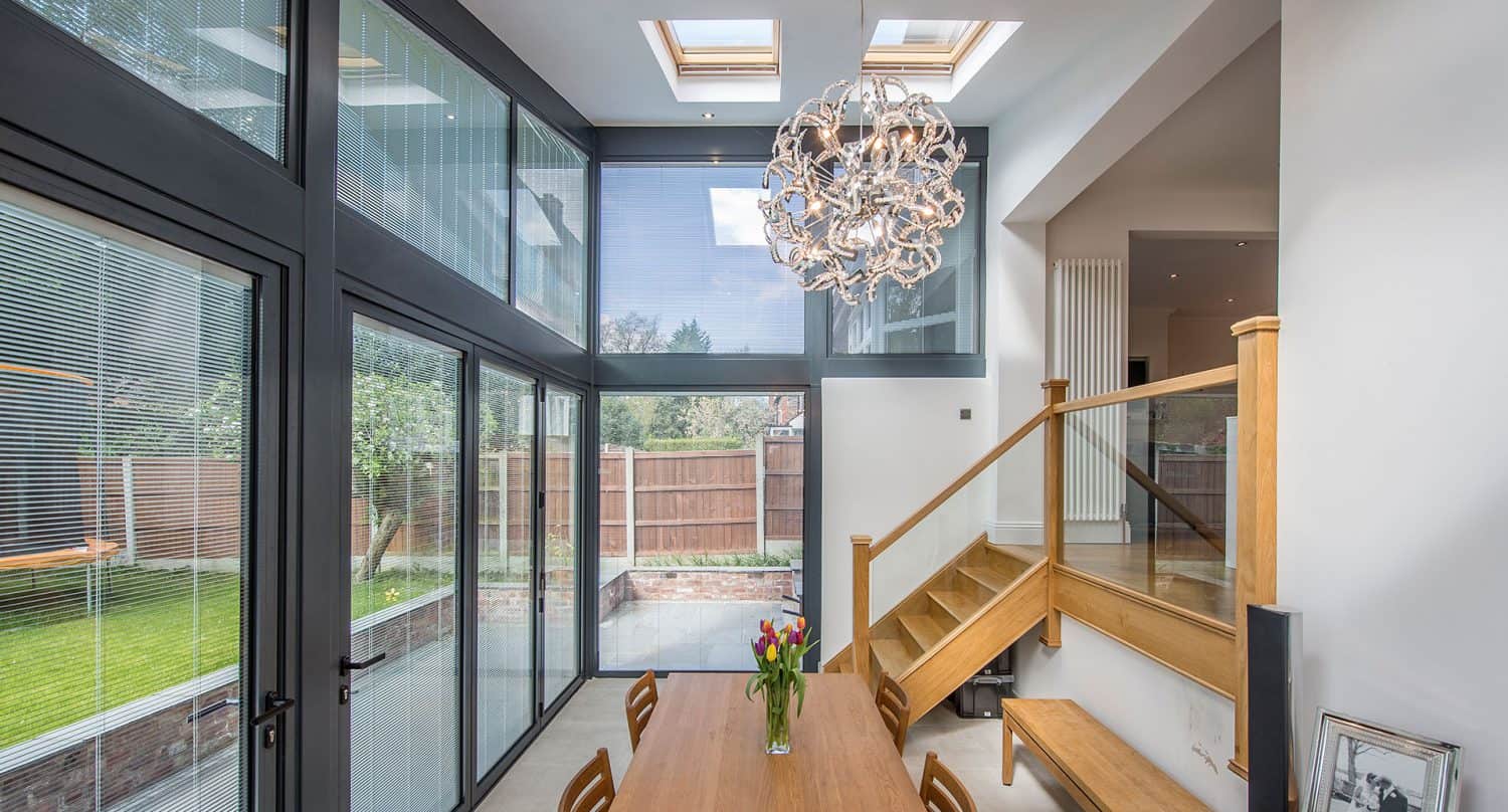 Modern dining area with large floor-to-ceiling windows, a wooden table with chairs, contemporary chandelier, wooden staircase, and view of a garden outside. Natural light fills the space through skylights and windows.