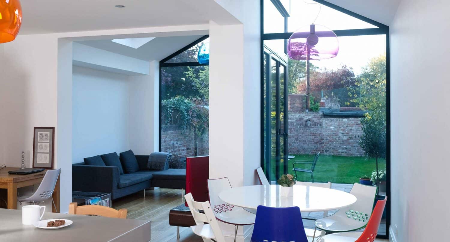 Modern living and dining area with large floor-to-ceiling windows, white walls, colorful chairs, a round dining table, gray sofa, wooden floor, and a view of a green garden and brick wall outside.