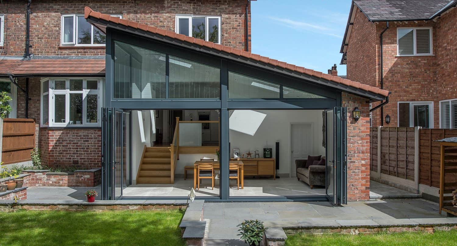 A modern house extension with large glass doors and windows, sloped roof, and brick walls, opening onto a patio and garden. Inside, there is a dining table, chairs, and a staircase leading upstairs.