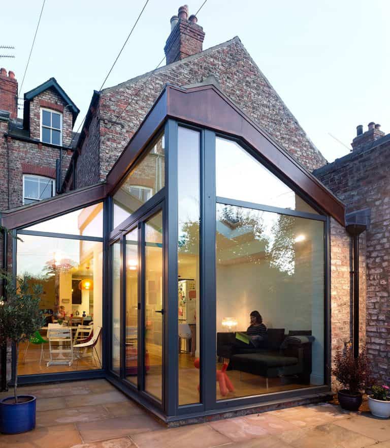 A modern glass extension with sharp angles is attached to a brick house. Inside, a person sits on a sofa, and a dining area with people is visible. Outdoor potted plants line the stone patio.