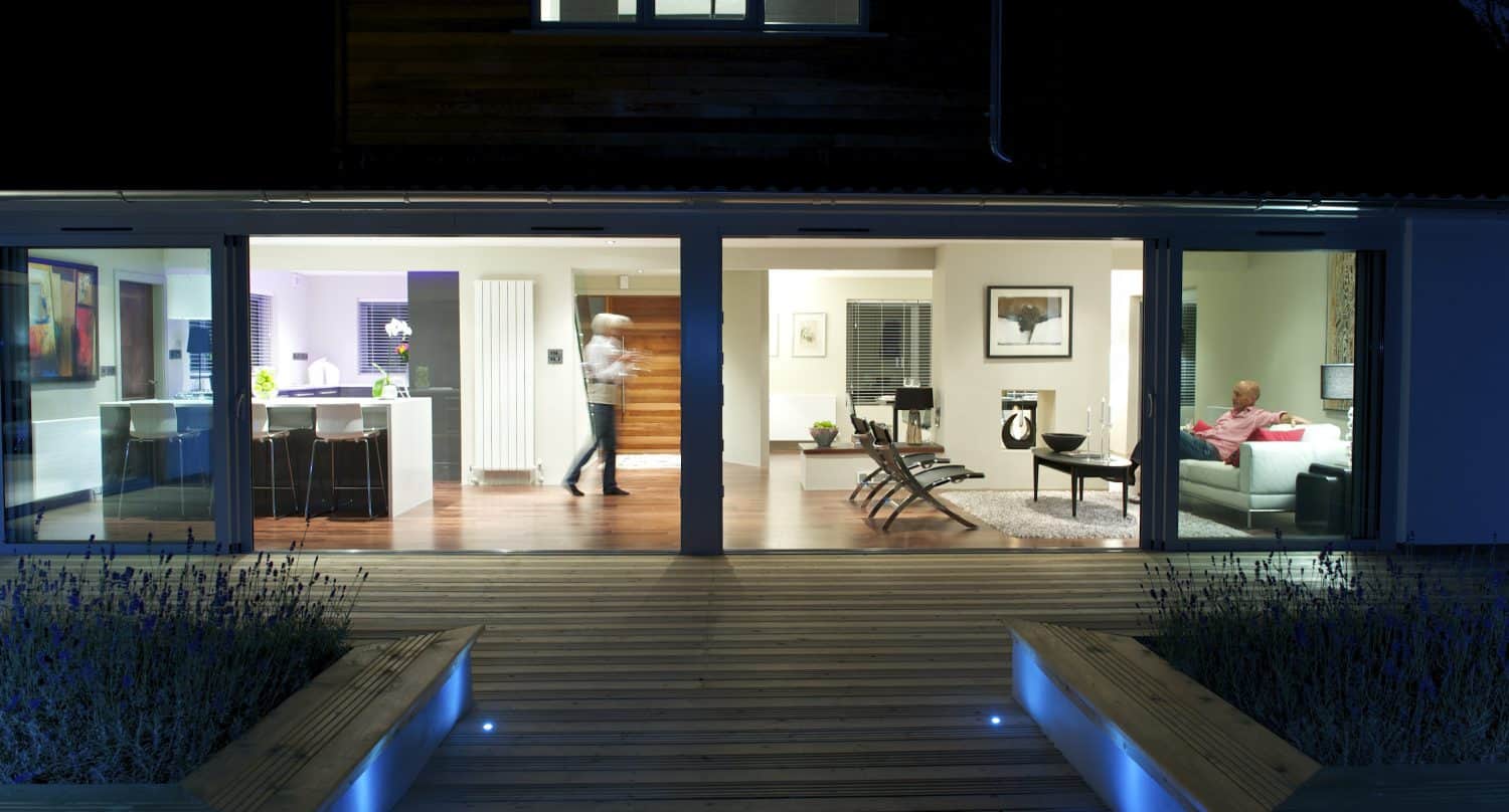 A modern house at night with large bifold doors open to a wooden deck; one person walks in the kitchen while another sits on a sofa in the living room, both spaces brightly lit inside.