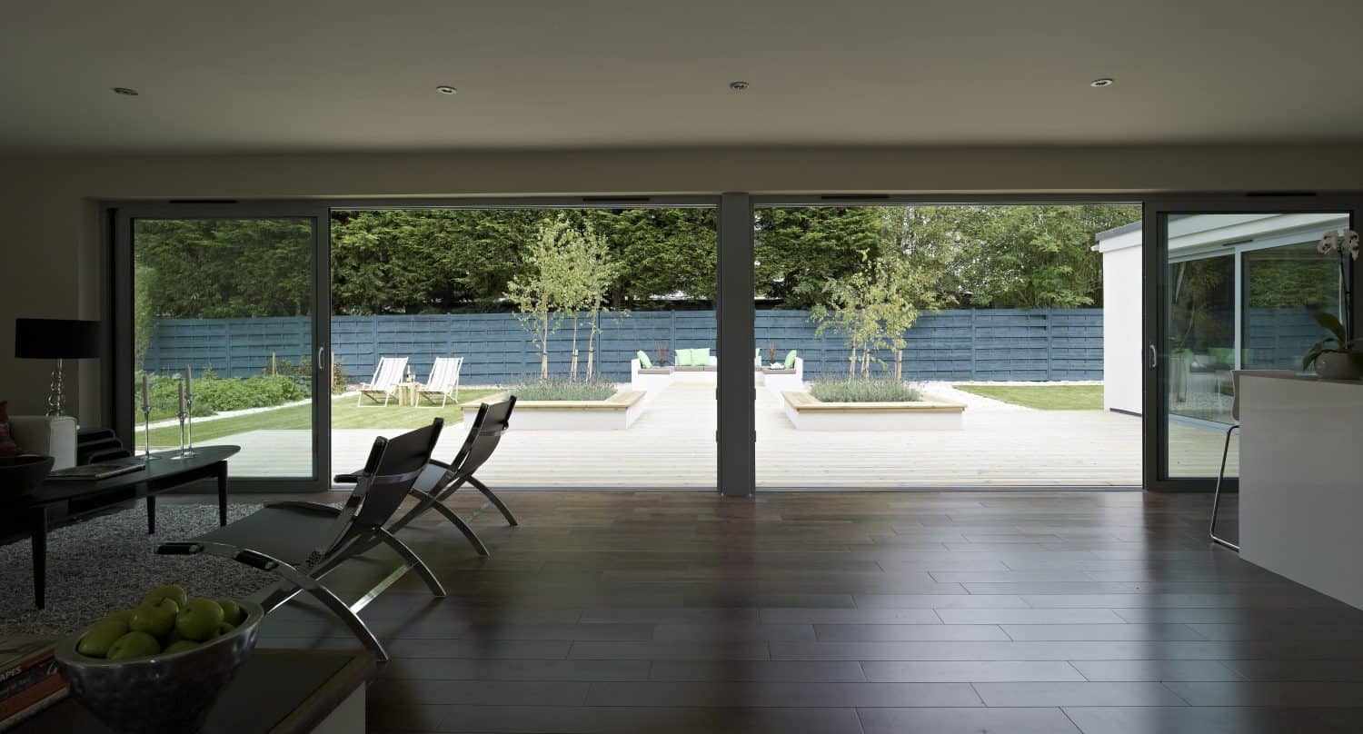 A modern living room with bespoke glass solutions features sliding glass doors that open to a patio and garden area with lounge chairs, potted trees, and a blue privacy fence in the background.