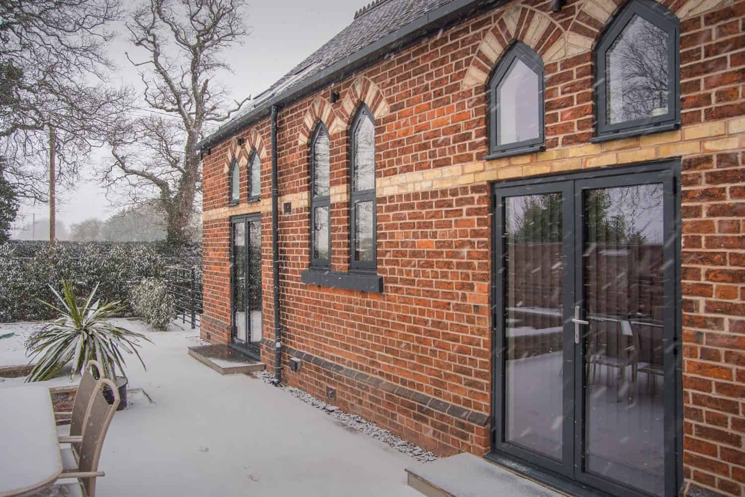 A red-brick building with arched windows and black-framed glass doors, surrounded by snow. An outdoor table and chairs sit on a snow-covered patio next to some trees and shrubs.