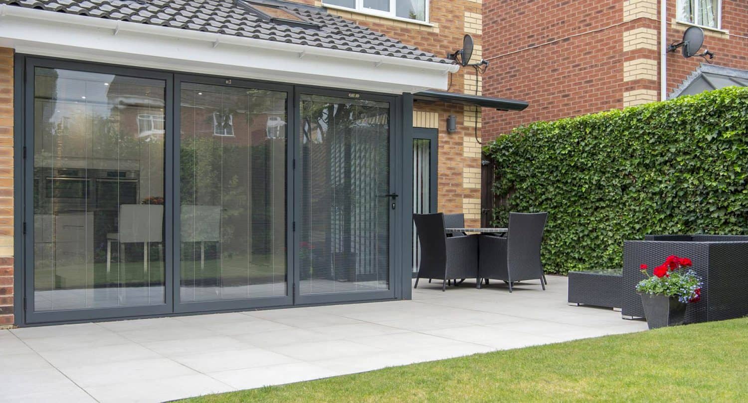 Modern patio with large glass folding doors, outdoor dining set, black wicker sofa, and potted red flowers. Brick house with hedges and neatly tiled patio floor visible.