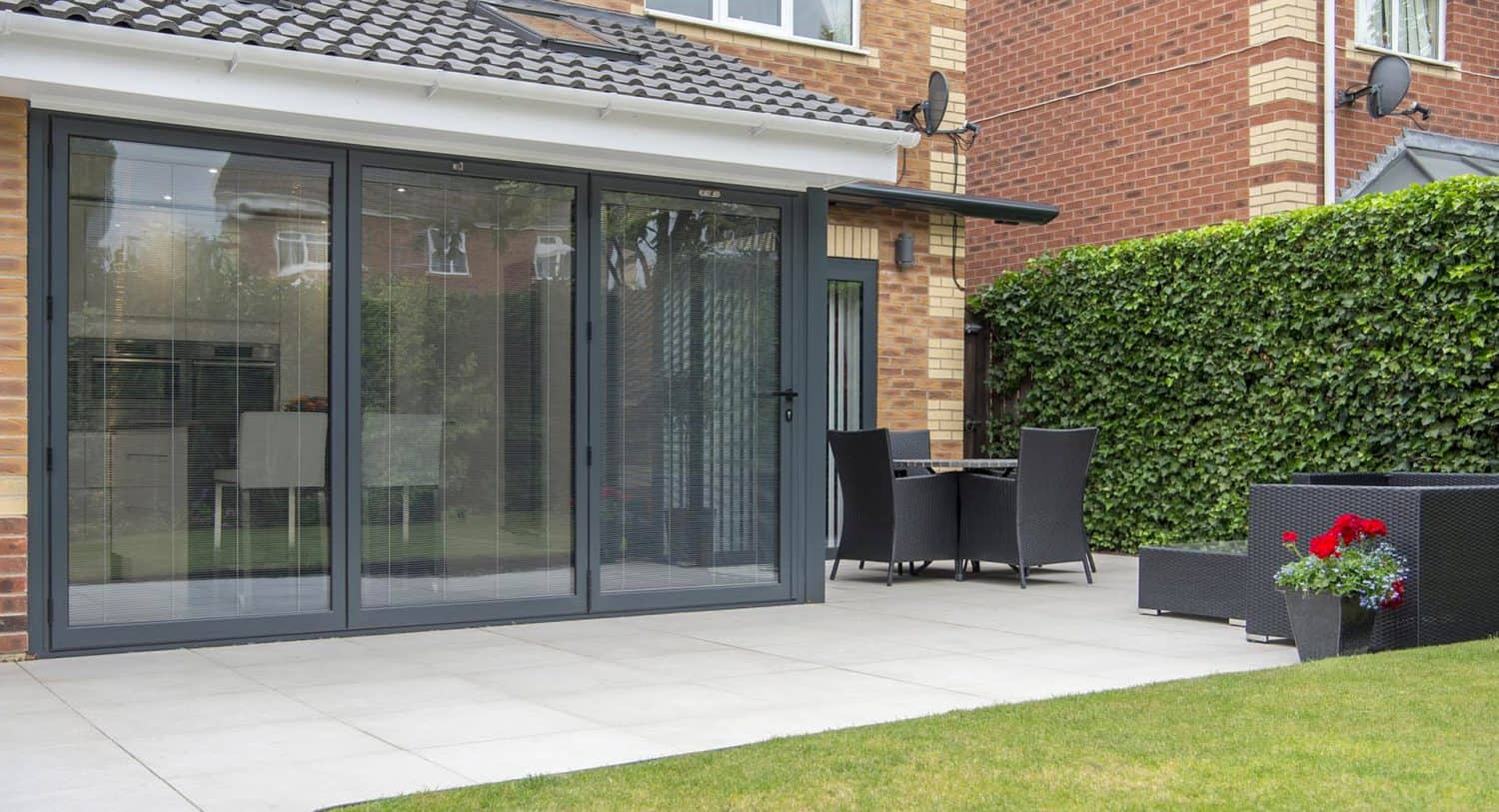 Modern patio with large glass folding doors, outdoor dining set, black wicker sofa, and potted red flowers. Brick house with hedges and neatly tiled patio floor visible.