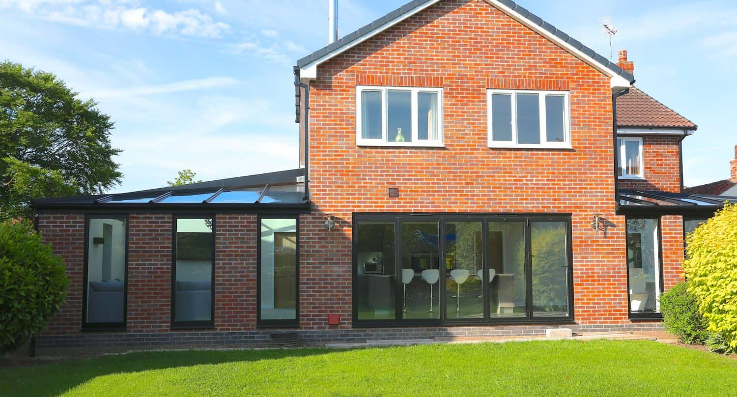 A modern brick house with large glass windows and doors, a flat-roofed extension, and a well-kept green lawn in the foreground, set under a clear blue sky.