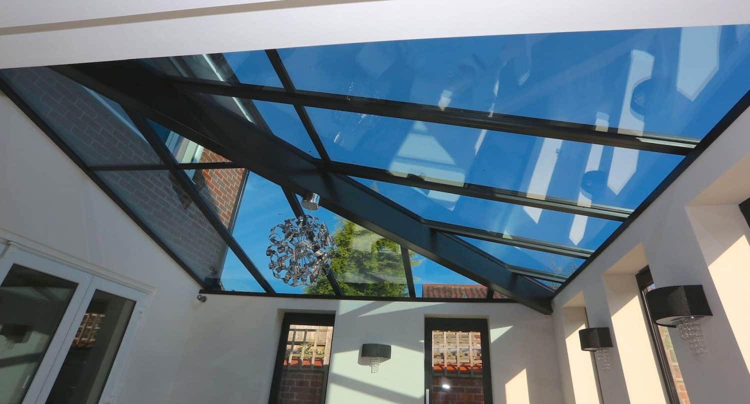 A modern glass roof with black metal frames lets natural light into a room. The blue sky and a brick building are visible through the glass. Inside, there is a decorative chandelier and wall lamps.