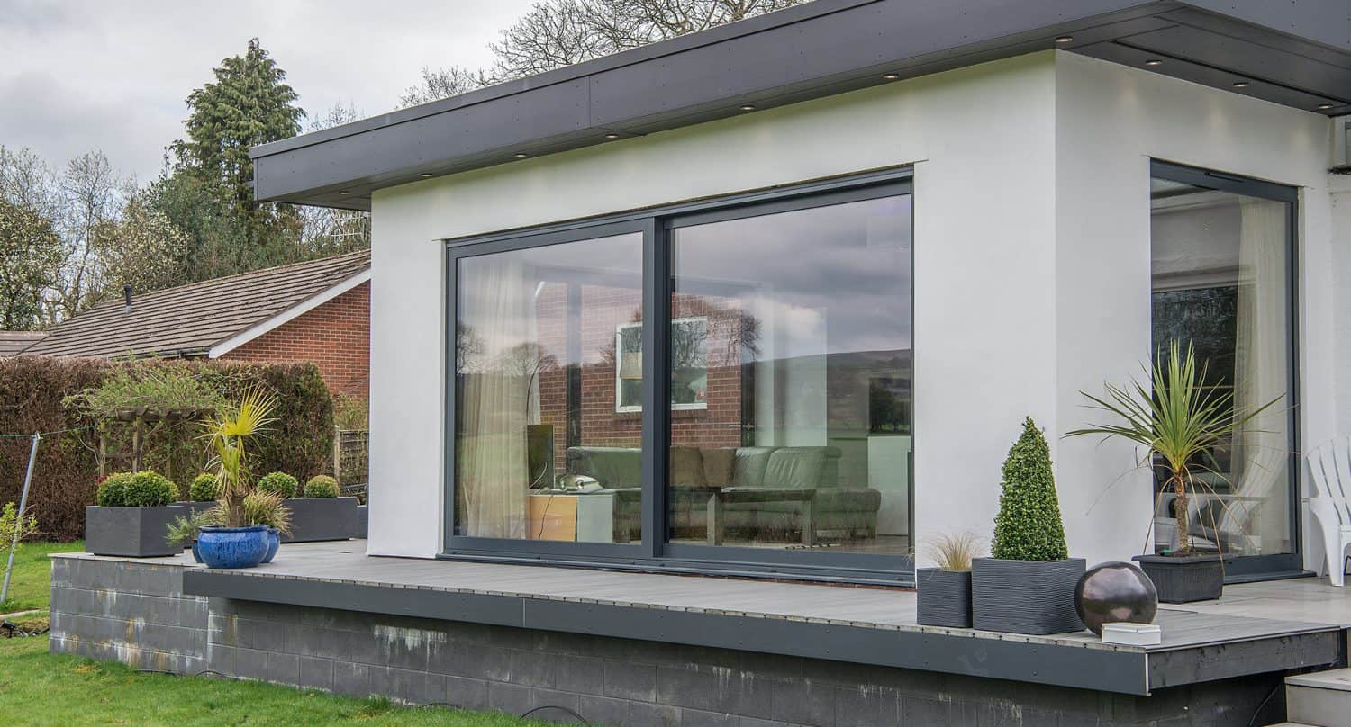 A modern house with large sliding glass doors, a white exterior, and a small patio decorated with various potted plants. The patio overlooks a green lawn and is surrounded by trees and neighboring homes.