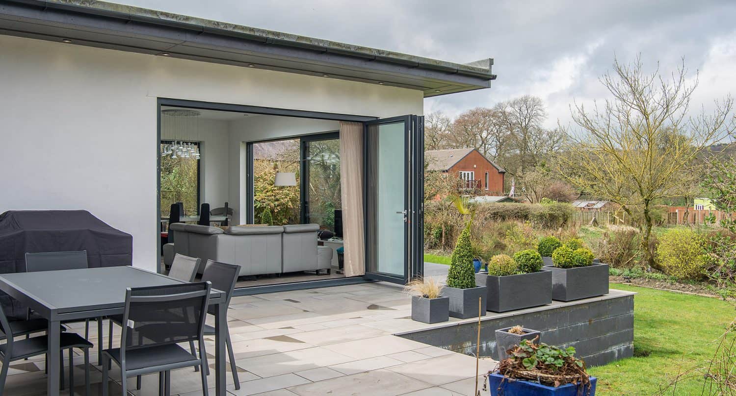 A modern patio with a dining table and chairs, a barbecue, and potted plants. Large glass doors open to a living room with a sofa, overlooking a garden with trees and a red brick house in the background.