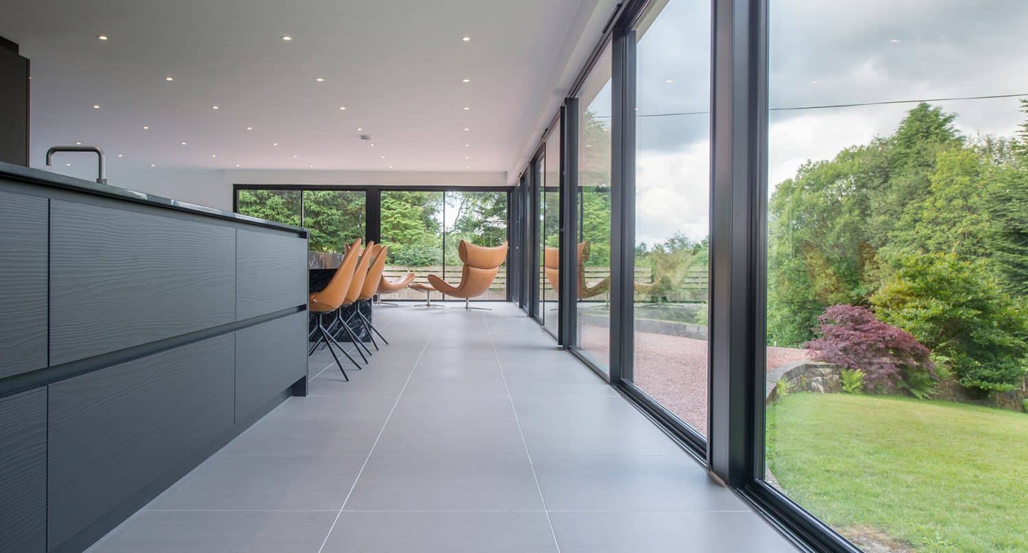 Modern kitchen and dining area with large floor-to-ceiling glass windows, gray tile flooring, and views of a lush green garden outside. Tan chairs surround the dining table, and two lounge chairs face the windows.