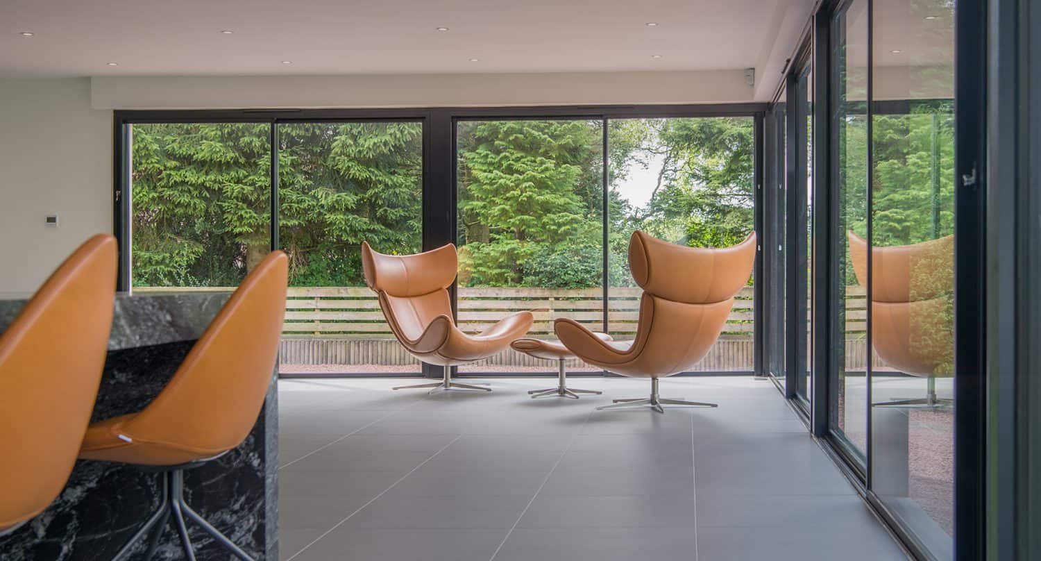 A modern room with large floor-to-ceiling windows, two orange swivel chairs facing each other, and a view of green trees outside. The floor is gray tile, and part of a marble counter is visible on the left.