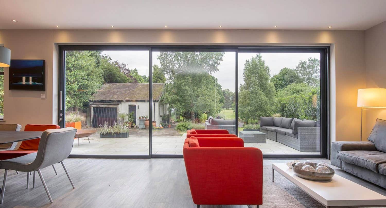 Modern living and dining area with red chairs, gray sofa, and large sliding glass doors opening to a spacious patio with outdoor seating, trees, and a garden shed in the background. Natural light brightens the room.